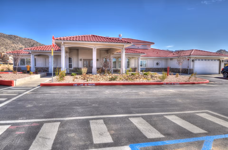 Front exterior view of a single-story building with a red tiled roof, beige walls, and a covered entrance supported by columns. There is a landscaped area with rocks and small plants in front of the building, a stop sign, and a parking lot with marked pedestrian crosswalks and fire lane no parking zones. Mountains are visible in the background under a clear blue sky.