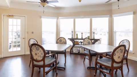Sunlit dining room with three tables, upholstered wooden chairs, and large bay windows with a glass door to the side.