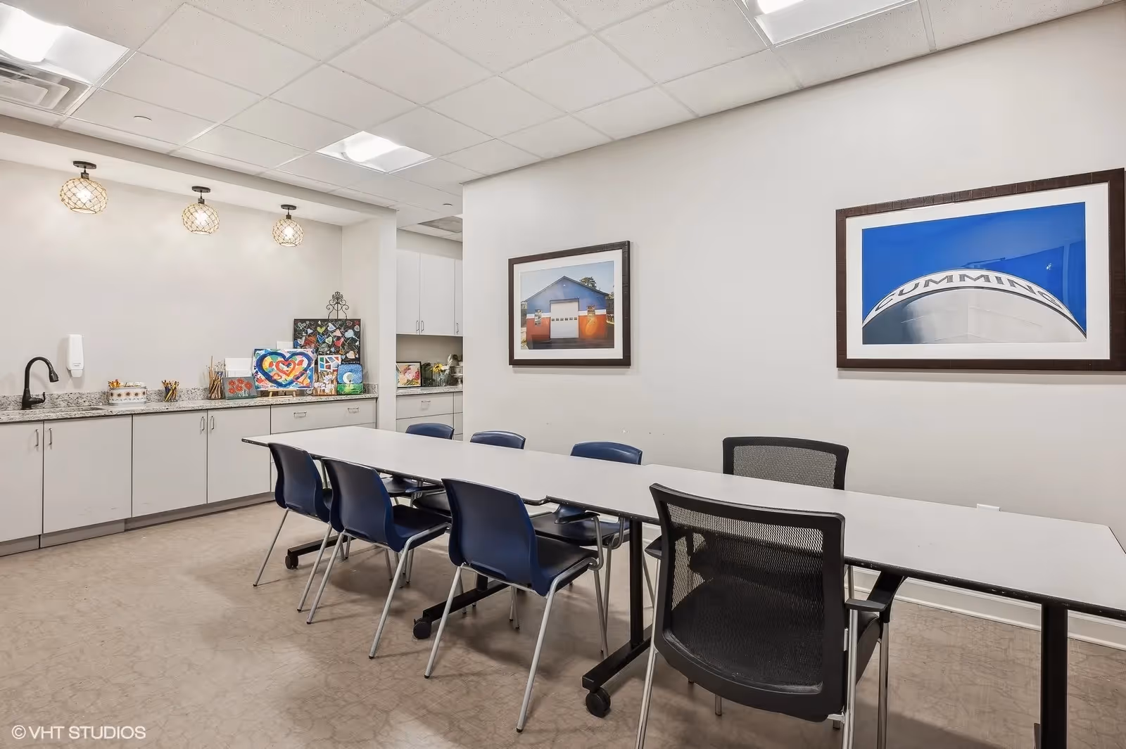 A well-lit interior room with a long white table surrounded by black and blue chairs. The back wall features white cabinets with a granite countertop, decorated with colorful artwork and craft supplies. Two framed photographs hang on the adjacent wall, and three pendant lights hang from the ceiling above the countertop.