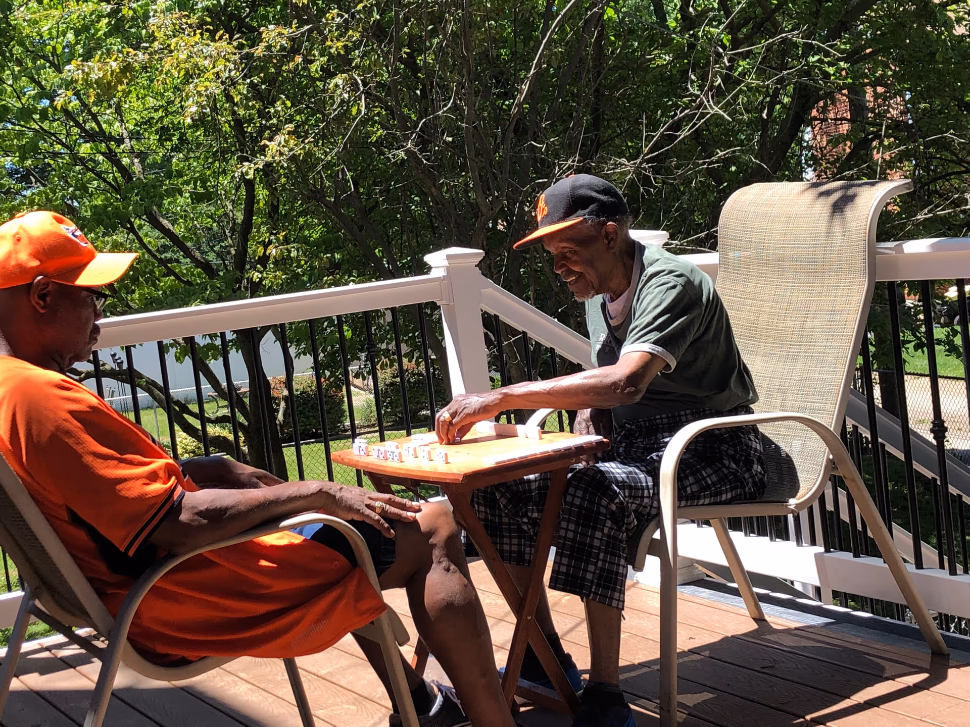 Two elderly men sitting on a wooden deck outdoors, playing a board game on a small table between them. One man is wearing an orange shirt and cap, while the other is wearing a dark green shirt and a black cap. Trees and greenery are visible in the background.
