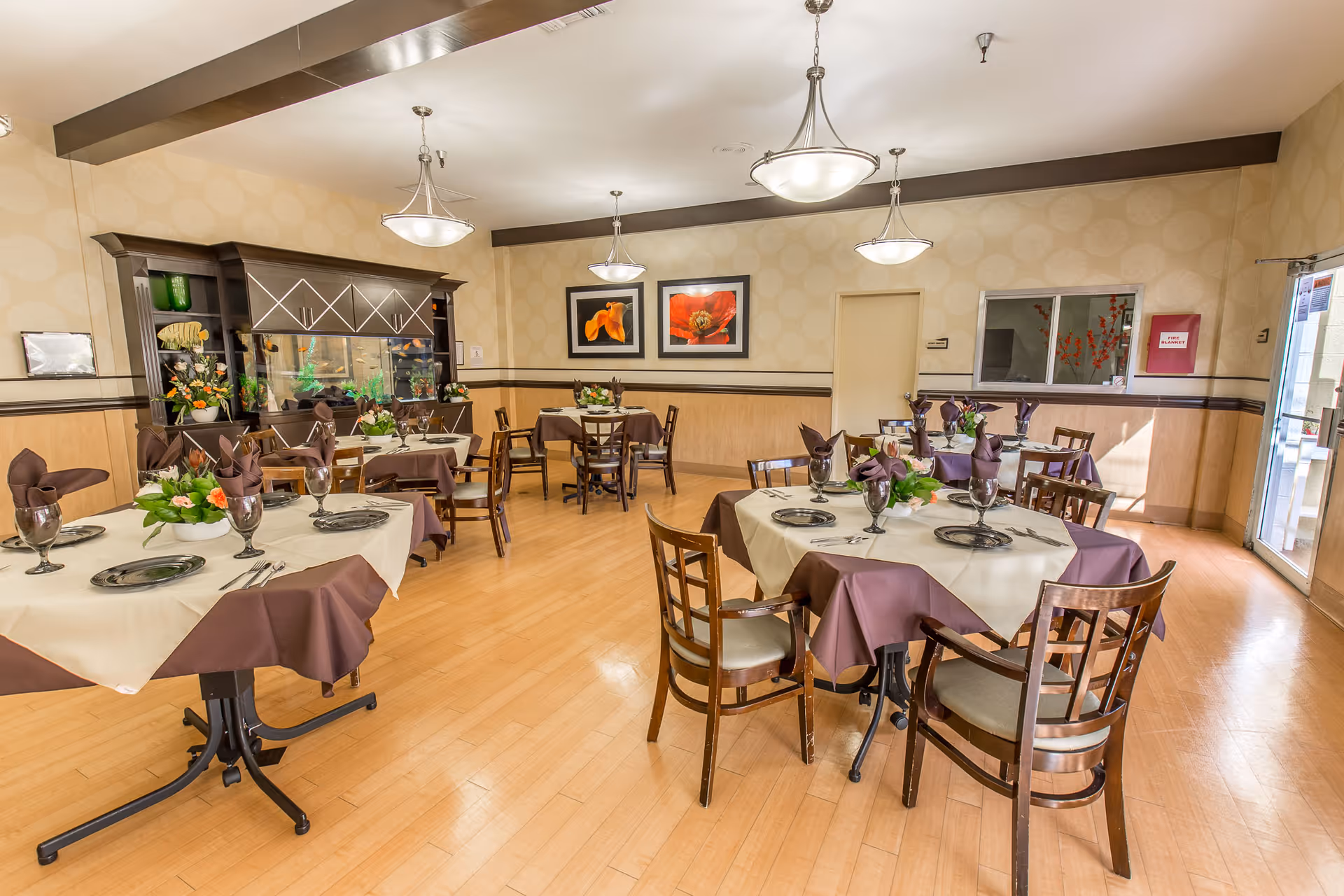 A dining room with several round tables covered with cream and brown tablecloths, each set with plates, glasses, silverware, and floral centerpieces. The room has wooden flooring, beige patterned walls with a dark trim, three hanging light fixtures, and framed floral artwork on the walls. There is a dark wooden cabinet with decorative items and plants, and a window and door on the right side letting in natural light.