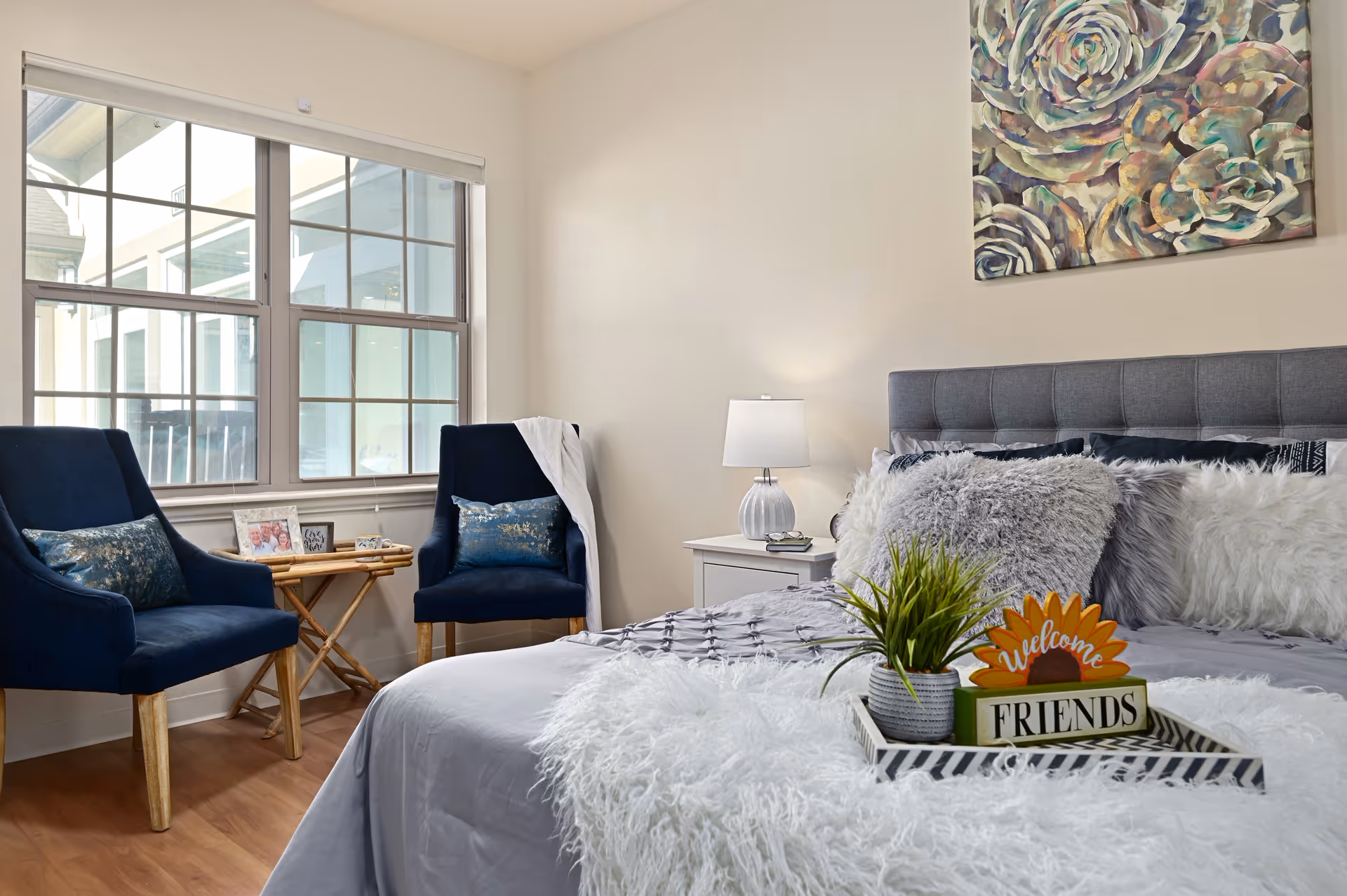 A cozy bedroom with a large bed adorned with fluffy pillows and a soft blanket. On the bed is a tray holding a small potted plant and a decorative sign that says 'Welcome Friends'. Two navy blue chairs with decorative pillows are positioned near a window with white blinds. A small wooden table between the chairs holds framed photos. A nightstand with a lamp is beside the bed, and a large floral painting hangs on the wall above the headboard.