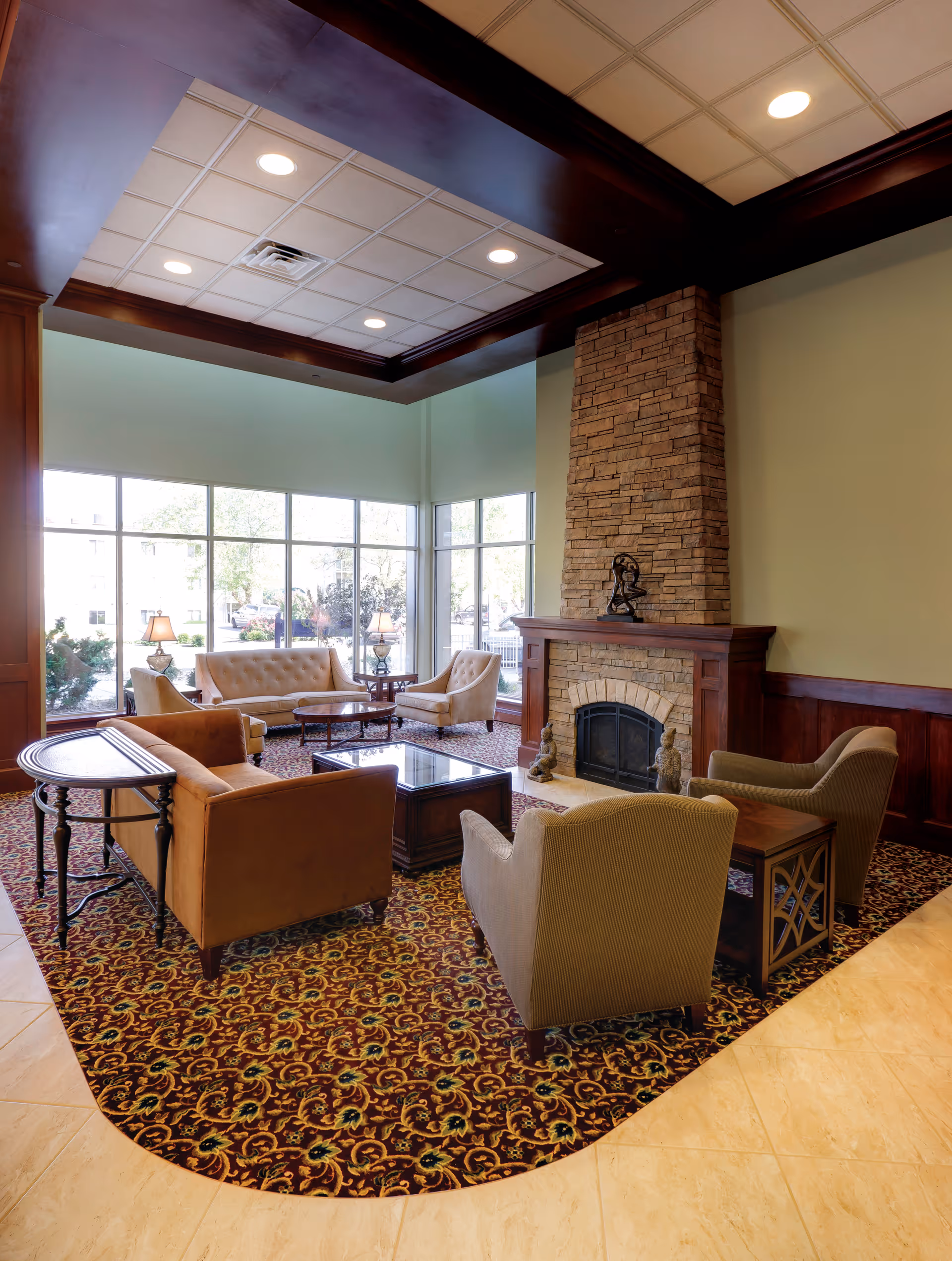A cozy seating area in a senior living facility featuring a stone fireplace with decorative sculptures on the mantel and hearth. The room has large windows letting in natural light, patterned carpet, and a mix of upholstered chairs and sofas arranged around a central coffee table. The ceiling has recessed lighting and dark wood trim.