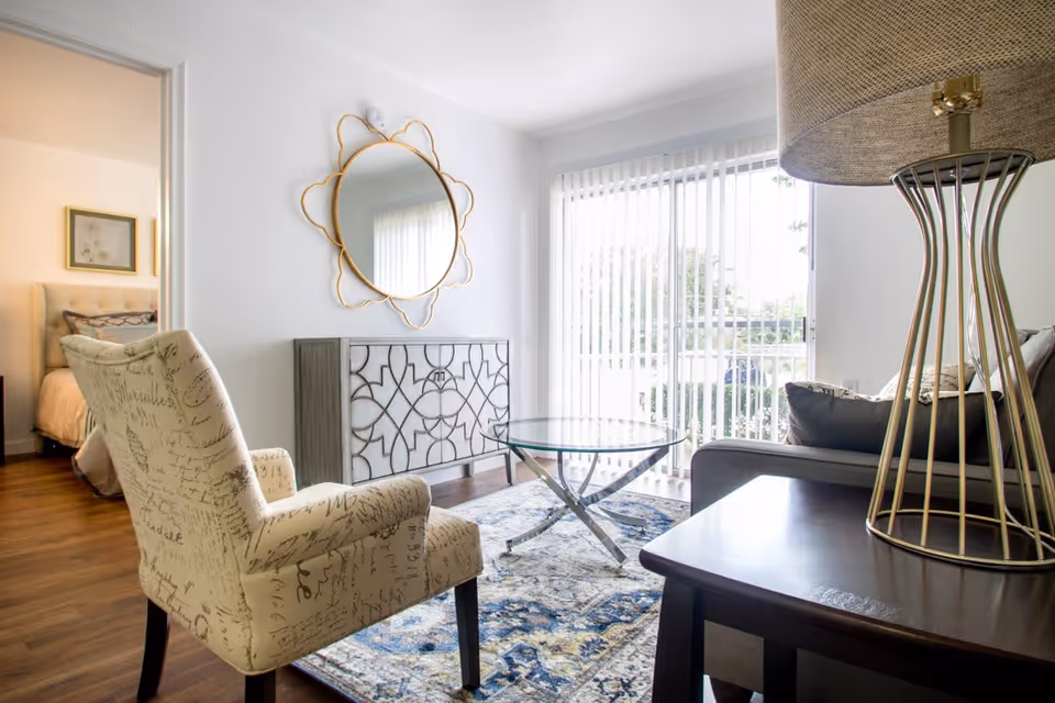 Bright living room with a patterned armchair, glass coffee table, sofa, decorative mirror, and sliding glass door to a balcony.