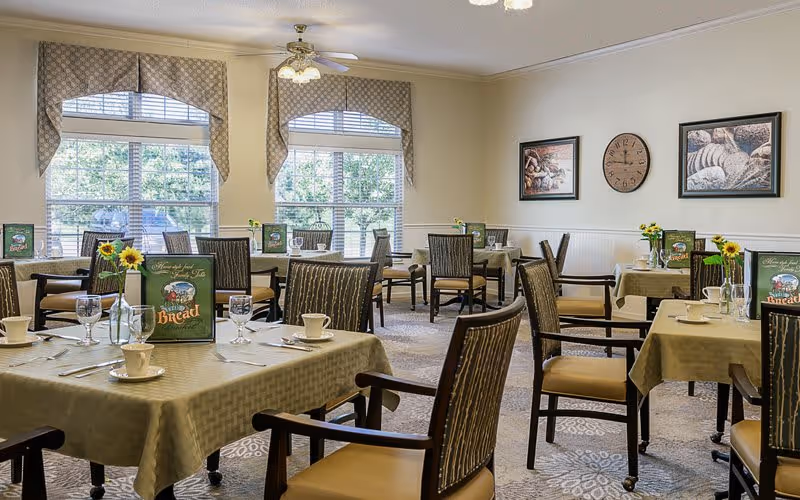 A dining room with several tables covered in beige tablecloths, each set with cups, glasses, utensils, and small vases holding sunflowers. The room has large windows with patterned valances letting in natural light, and the walls are decorated with framed pictures and a clock.