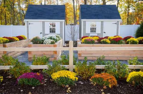 Two small white sheds with gray roofs behind a white fence, surrounded by colorful flower beds.