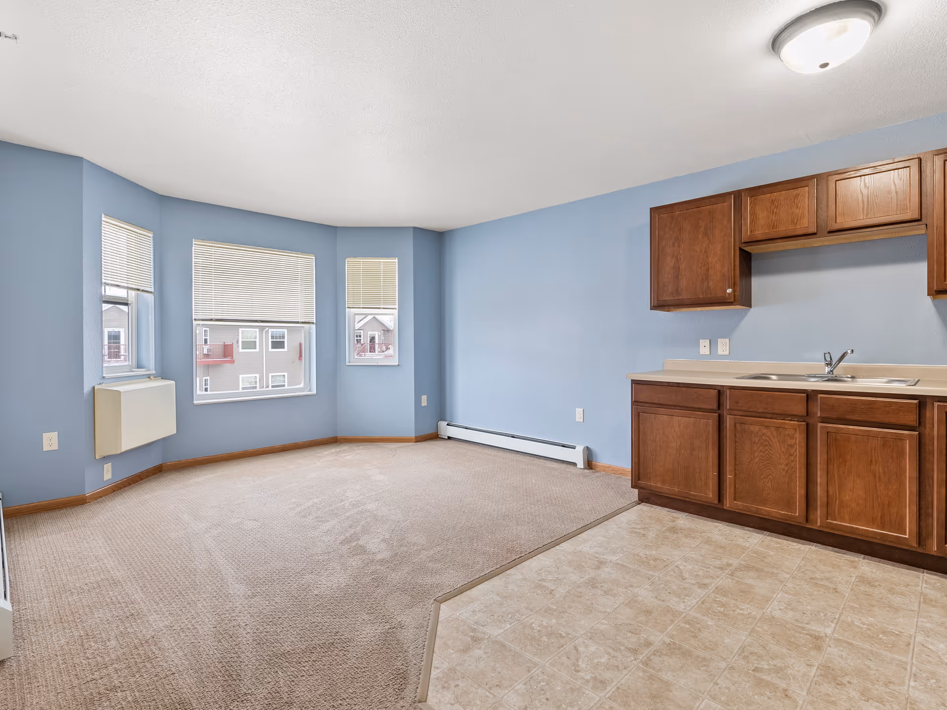 Empty room with light blue walls, beige carpet, and three windows with blinds. The room has a small kitchen area with wooden cabinets, a countertop, and a sink. The floor near the kitchen is tiled.