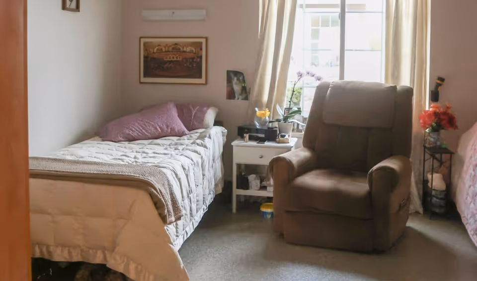 A cozy bedroom shows a single bed, beige recliner, nightstand by a window.