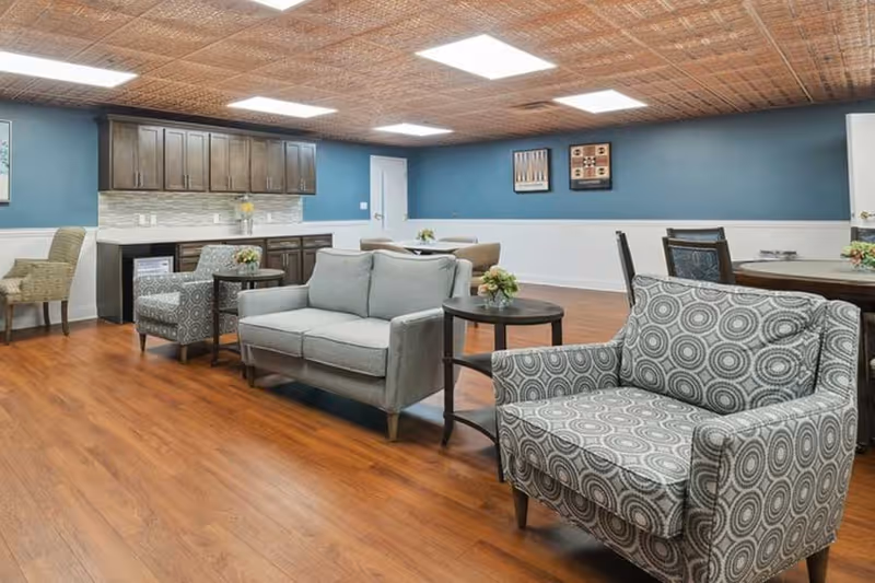 A senior living facility common area with wooden flooring, a blue accent wall, and a ceiling with square recessed lights. The room features a variety of seating options including a patterned armchair, a gray loveseat, and other chairs around tables. There are cabinets and a countertop along one wall, with small floral arrangements on the tables.