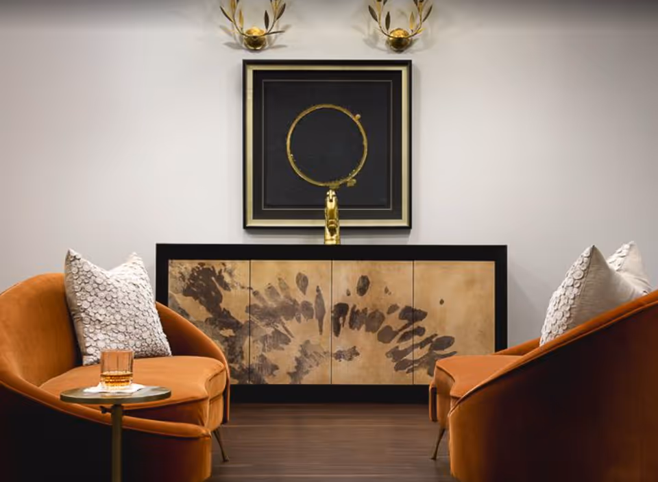 Two orange curved lounge chairs face a decorative console with framed art, gold wall sconces, and a small side table holding a glass.