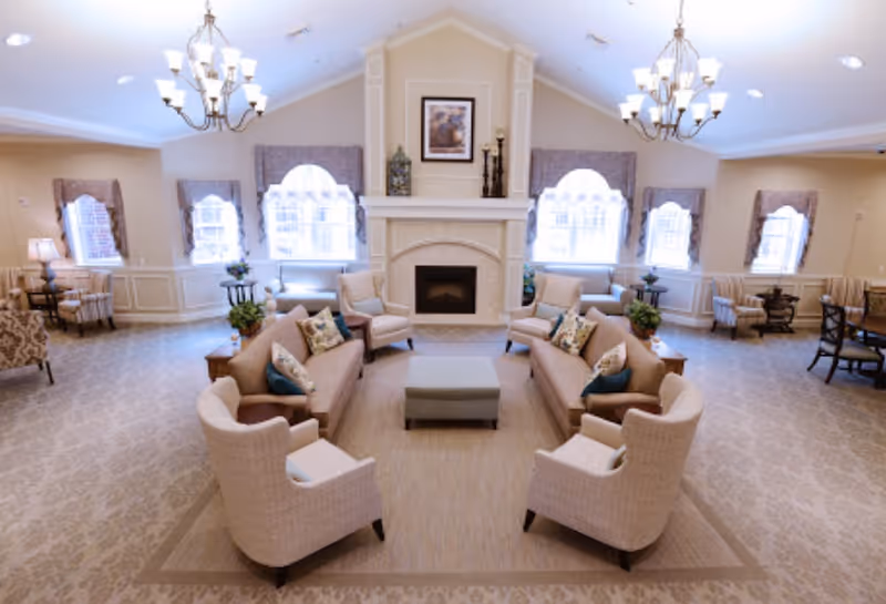 A spacious and well-lit living room area with beige walls and carpet. The room features two beige sofas facing each other with patterned pillows, four matching armchairs, and a central ottoman. There is a fireplace with decorative items and a framed picture above it. Multiple windows with valances allow natural light to fill the room. Two chandeliers hang from the ceiling, and additional seating areas with chairs and small tables are visible along the walls.