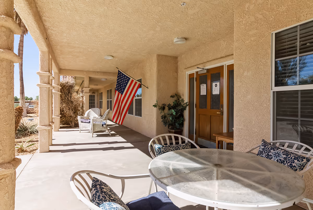Covered outdoor patio area with a round glass table and chairs with cushions, an American flag hanging on the wall, potted plants, and a wooden door with windows. The patio has textured beige walls and columns, with sunlight casting shadows on the floor.