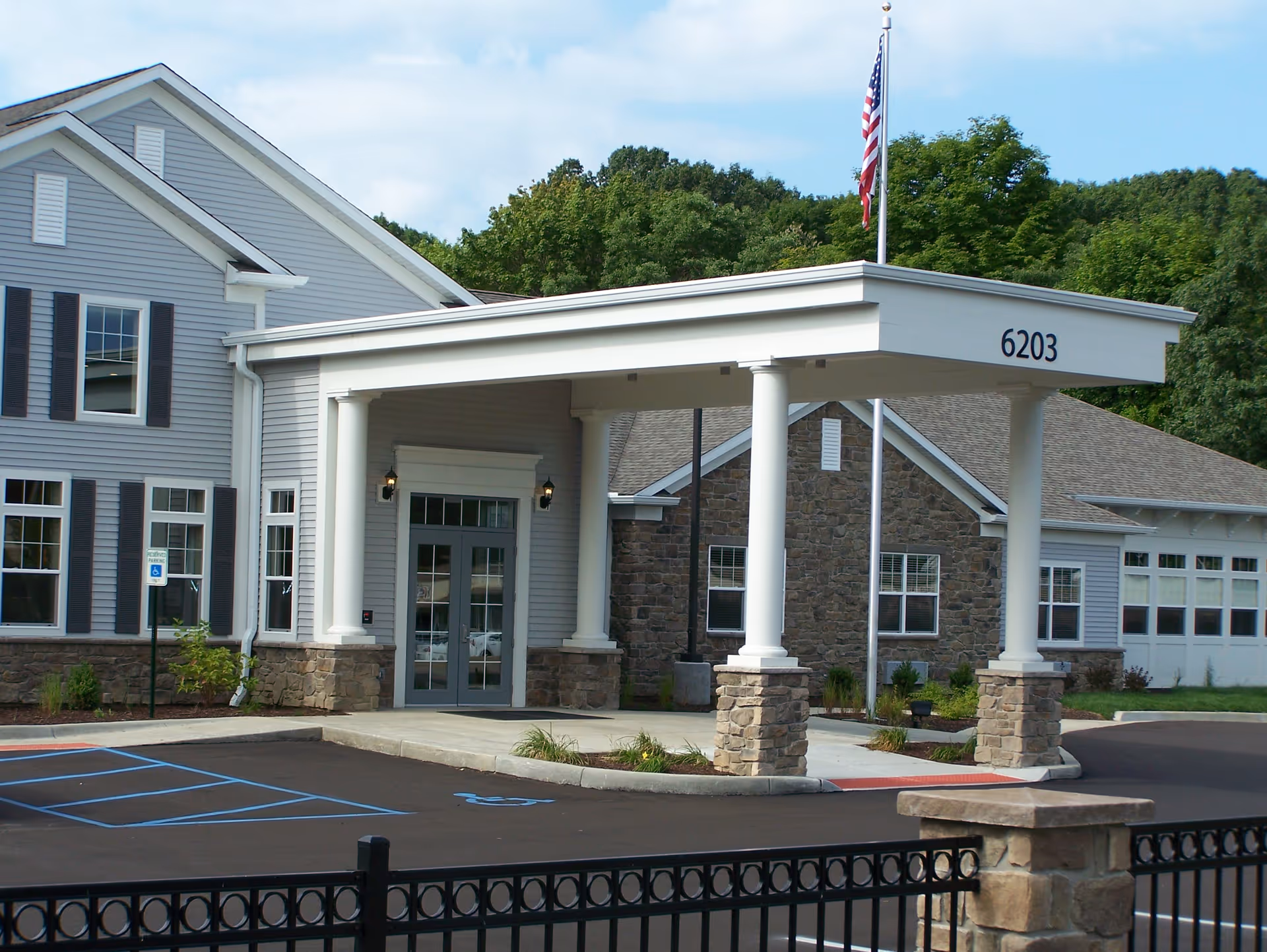Entrance of a senior living facility with a covered drop-off area supported by white columns with stone bases. The building has gray siding and stone accents, with an American flag flying on a flagpole. There are handicap parking spaces marked in blue in front of the entrance.
