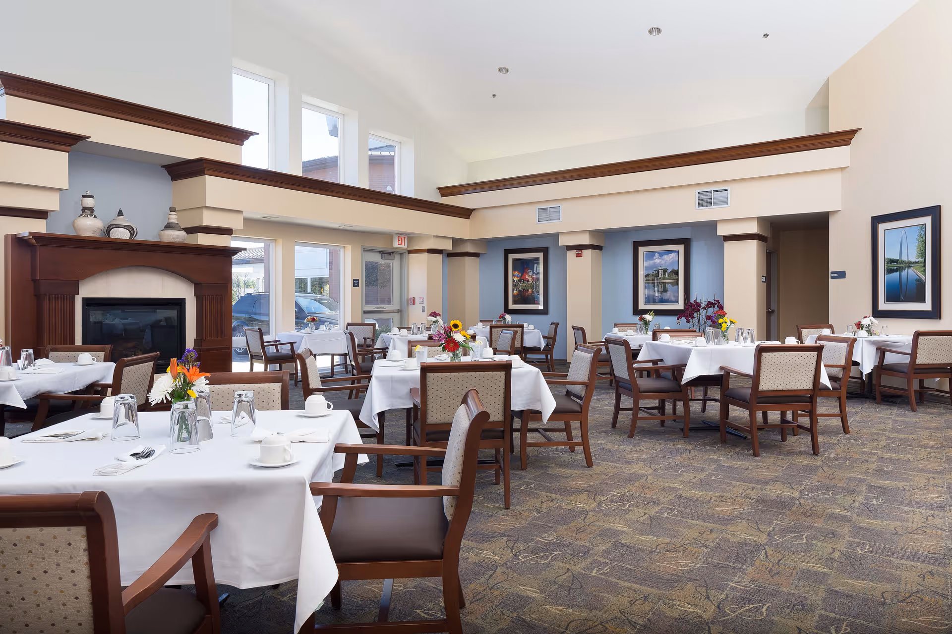 A spacious dining room with multiple tables covered in white tablecloths, each set with cups, glasses, and silverware. The room features wooden chairs with cushioned seats, a fireplace with decorative vases on the mantel, large windows letting in natural light, and framed artwork on the walls.