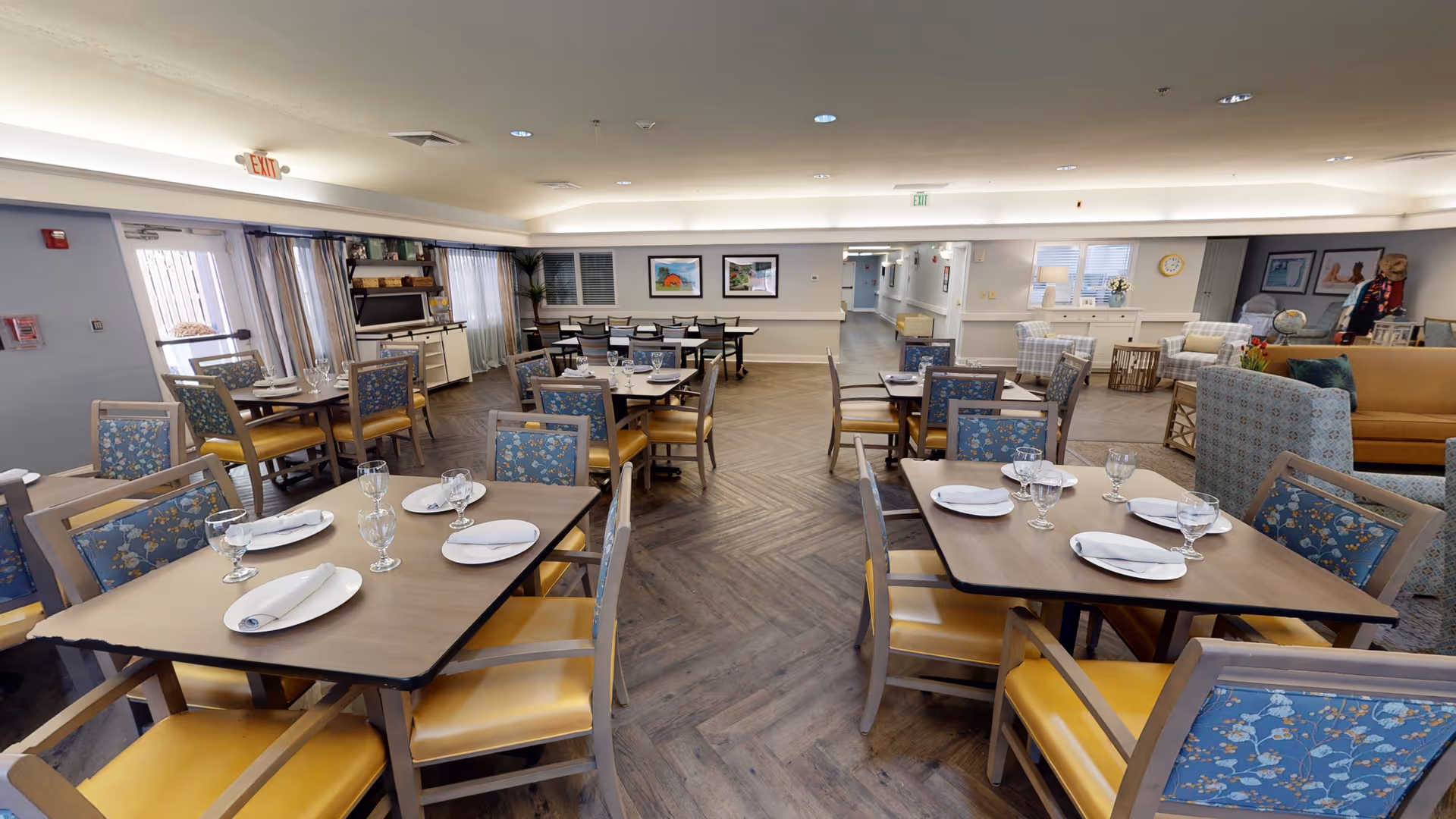Spacious dining room with multiple set tables and patterned cushioned chairs in a senior living facility.