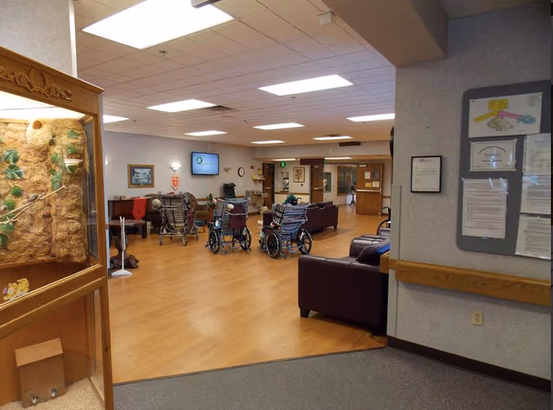 A senior living common area with several wheelchairs facing couches and a wall-mounted TV, bulletin boards, and wood-look flooring.