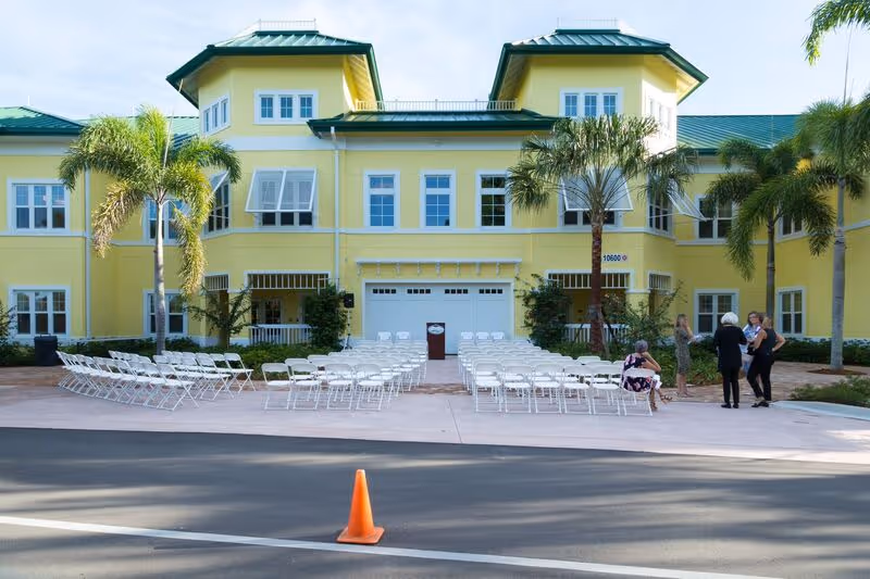 Outdoor view of a yellow two-story building with green roofs, palm trees, and rows of white folding chairs arranged facing a podium, with a few people standing and sitting nearby on a paved area.