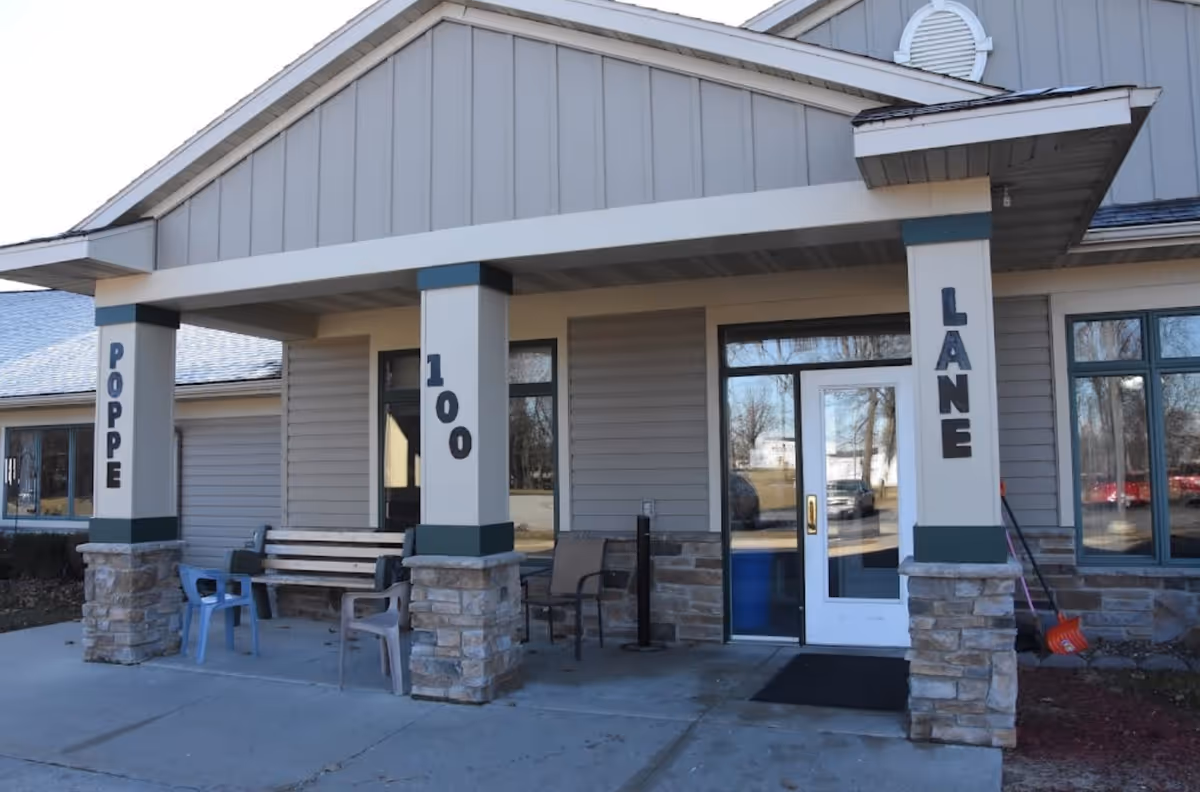 Entrance of a building with a covered porch supported by three columns. The columns have the words 'POPPE', '100', and 'LANE' vertically displayed. There are several chairs and a bench on the porch, and a glass door entrance is visible.