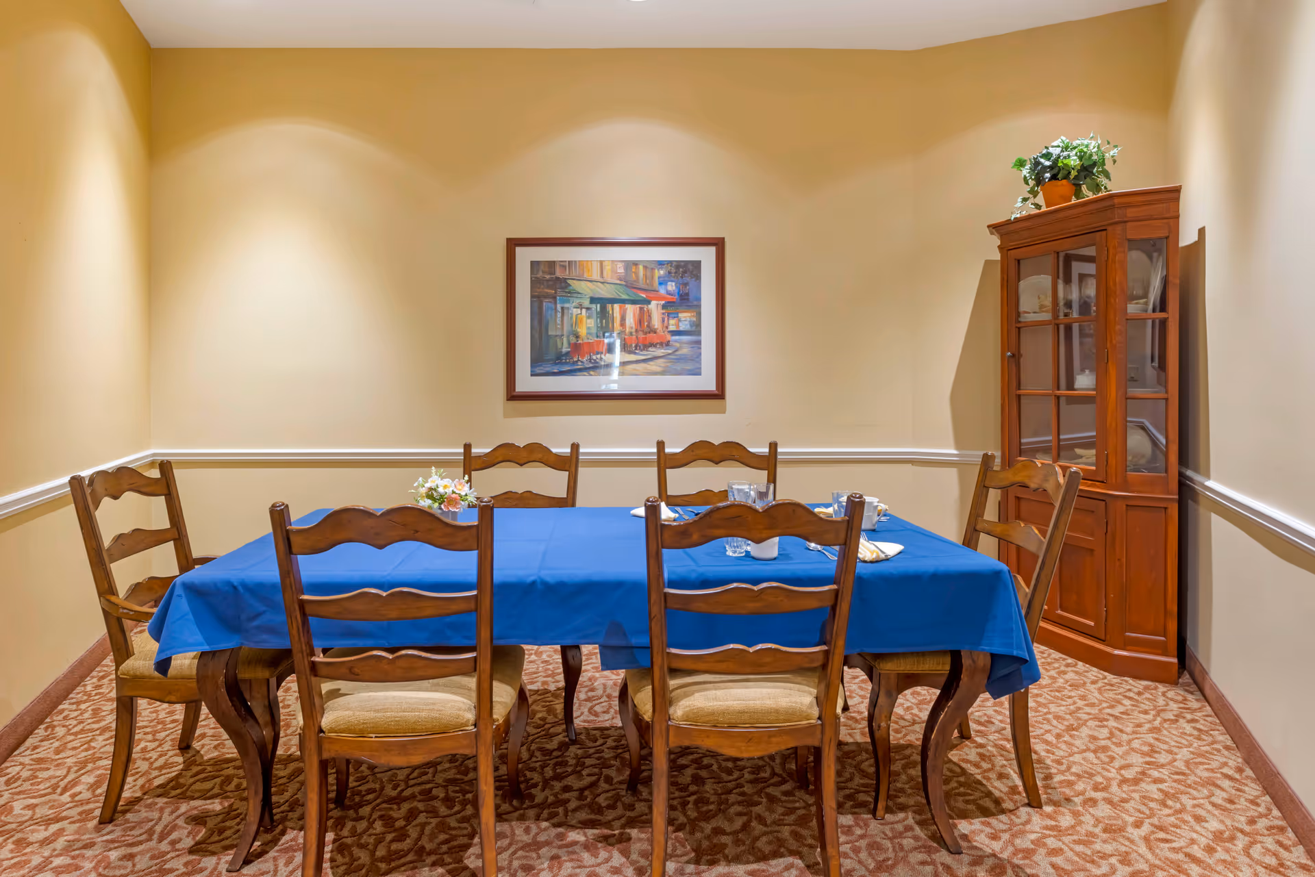 A dining room with a rectangular wooden table covered with a blue tablecloth and six wooden chairs with cushioned seats. On the table are some small decorative items and place settings. The room has beige walls with white trim, a patterned carpet, a framed painting on the wall, and a wooden corner cabinet with a potted plant on top.