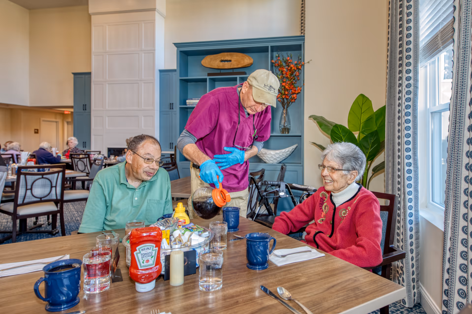 An elderly man and woman sit at a dining table in a bright dining room while a staff member wearing a purple shirt, beige cap, and blue gloves pours coffee into a mug. The table has condiments, glasses of water, and blue mugs. Other elderly people are seated at tables in the background. The room has large windows with patterned curtains and blue cabinetry with decorative items.