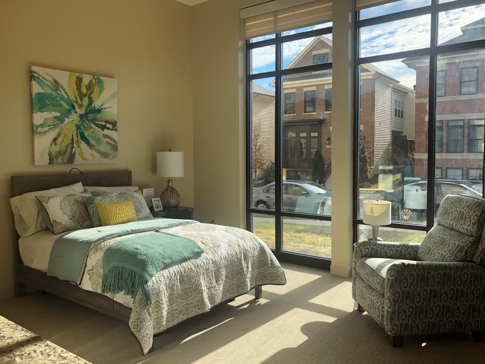 A bright bedroom with a large window showing a view of neighboring houses and parked cars. The room features a bed with patterned bedding and multiple pillows, a bedside table with a lamp and picture frame, and a patterned armchair with a small lamp beside it.