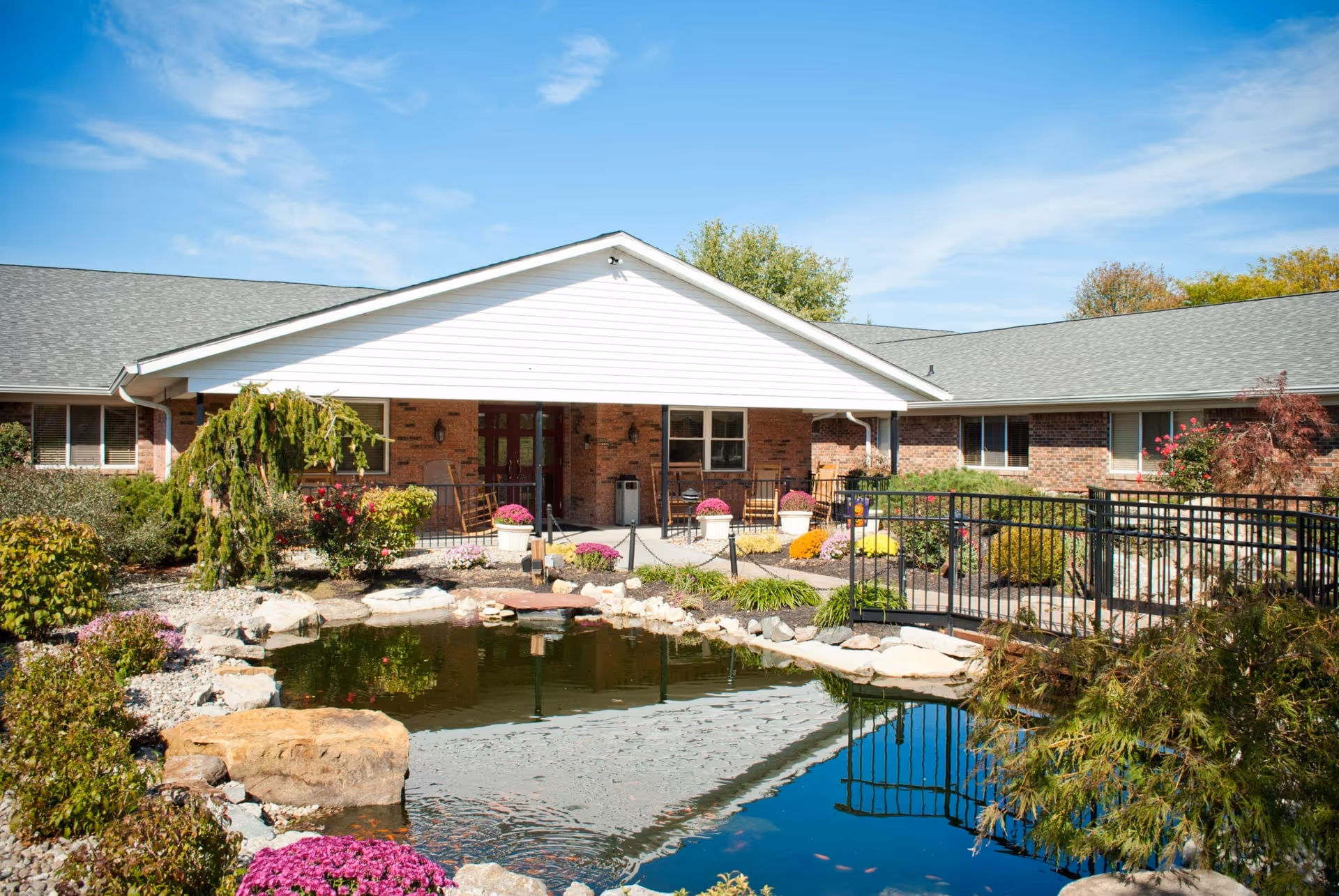 Outdoor view of Arbor Grove Village featuring a landscaped garden with a pond, rocks, colorful flowers, and a small black metal bridge. The building has a brick exterior with a white gable roof and a covered porch area with chairs and potted plants.