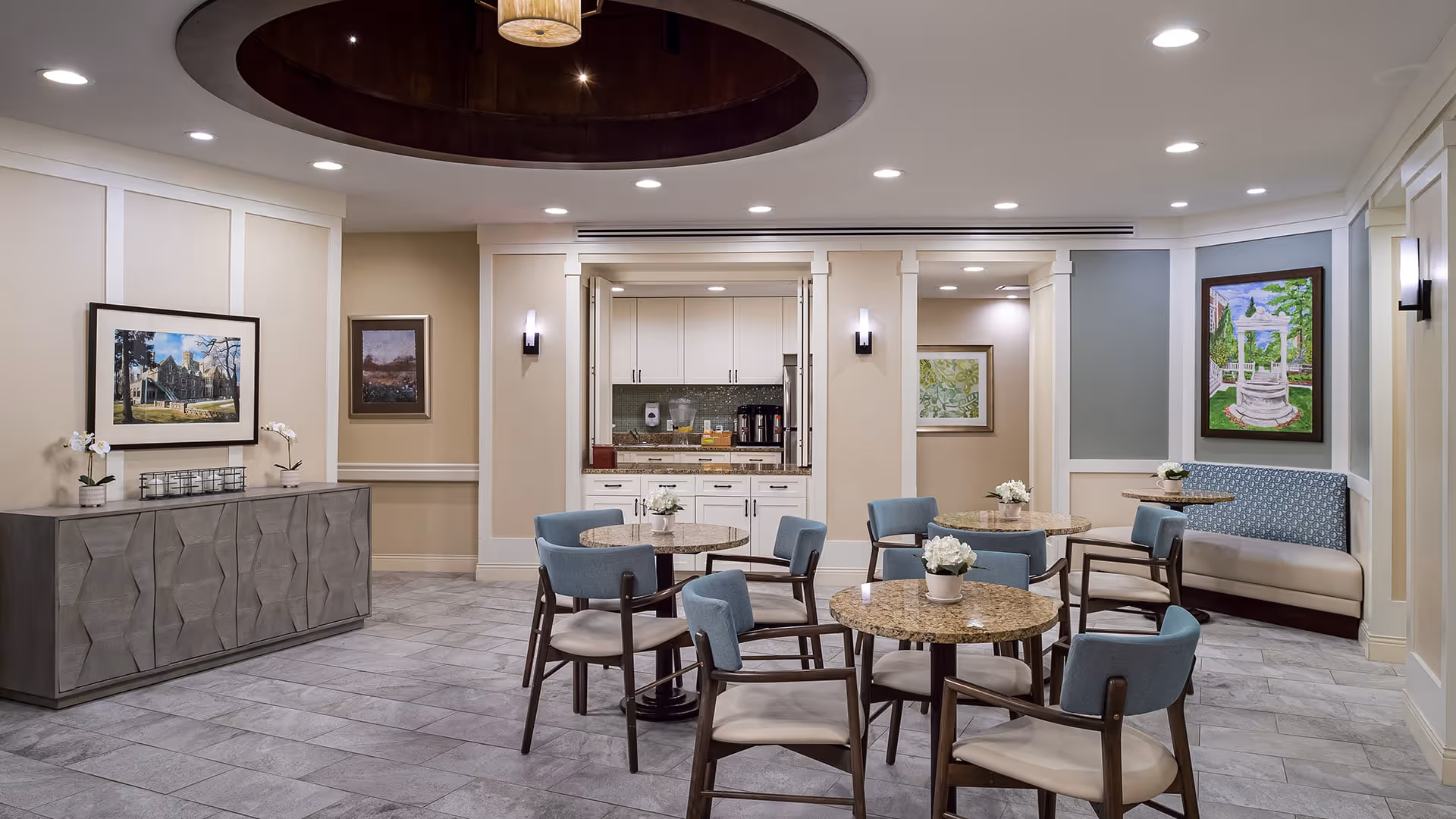 A well-lit common area with round tables and blue cushioned chairs arranged around them. There is a built-in kitchenette with white cabinets and a coffee machine in the background. The room features light-colored walls adorned with framed artwork and a decorative ceiling light fixture. A cushioned bench with a patterned backrest is positioned against the wall on the right side.