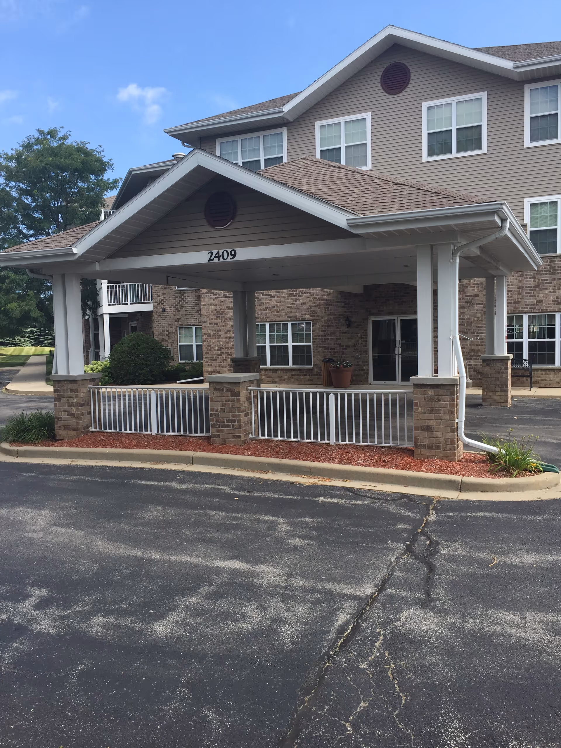 Entrance area of a senior living facility with a covered drop-off zone supported by white columns and brick bases. The building has beige siding and multiple windows, with the number 2409 displayed on the front of the covered area. There is a paved driveway and some landscaping with mulch and plants around the entrance.