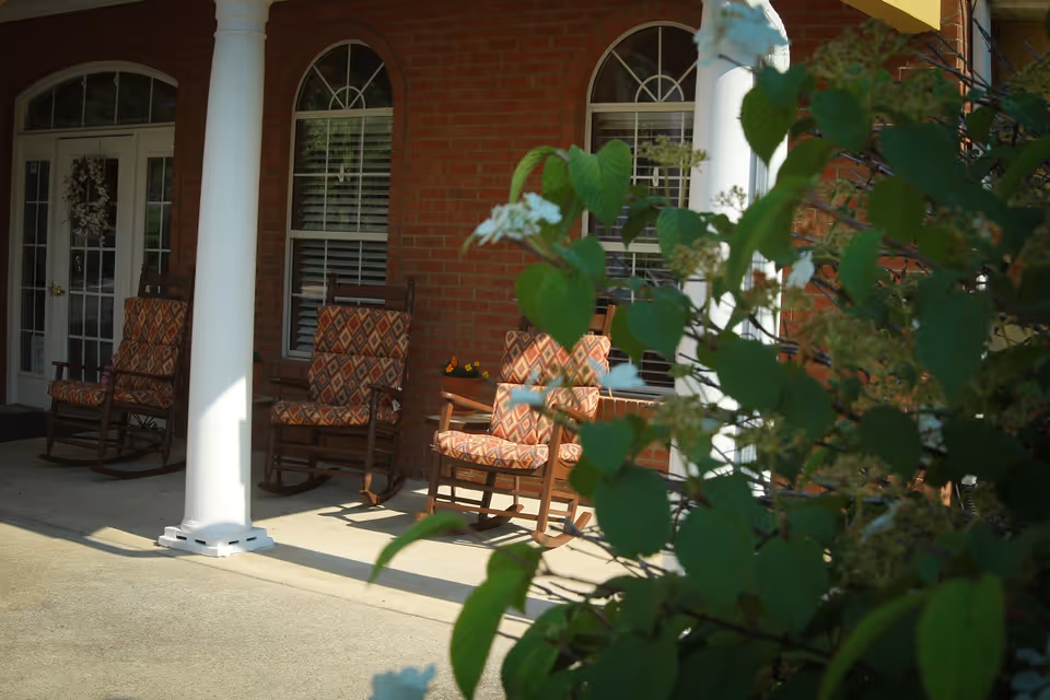 Front porch of a brick building with white columns and patterned rocking chairs, partially obscured by flowering shrubs.