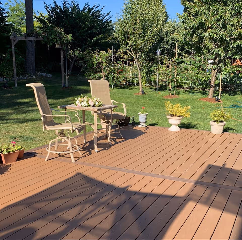 A wooden deck with two high-back chairs and a small table topped with flowers, surrounded by potted plants and a grassy backyard with trees.