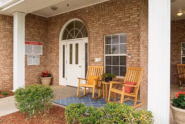 Front porch area of a brick building with a white door featuring glass panels and an arched window above. Two wooden rocking chairs with cushions sit on a blue patterned rug next to a small table with a potted plant. There are white columns supporting the porch roof and green shrubs planted in mulch beds in front.