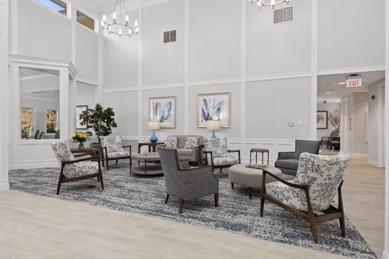 A spacious and well-lit living room area with high ceilings, featuring a mix of patterned and solid armchairs arranged around a round coffee table on a large blue and gray patterned rug. The walls are light gray with white trim, decorated with two framed botanical prints and two table lamps with blue bases. There is a large mirror on the left wall and an exit sign visible in the hallway on the right.