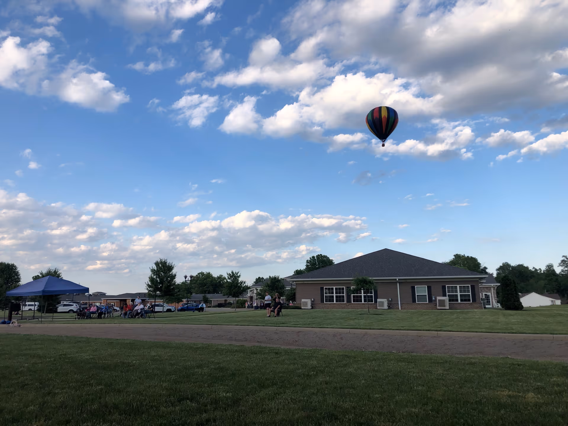 Single-story brick senior living building with people gathered on the lawn under a canopy and a hot air balloon in the sky.