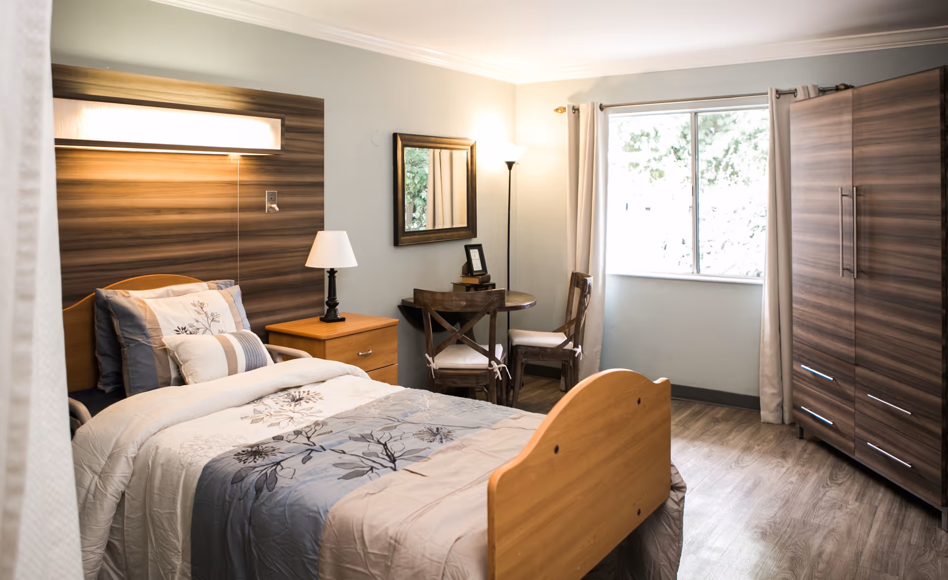 A well-lit nursing home bedroom with a single bed featuring floral patterned bedding. Next to the bed is a wooden nightstand with a lamp. There is a small round table with two wooden chairs near a window with beige curtains. A large wooden wardrobe is positioned against the wall, and a mirror hangs above the table. The room has light-colored walls and wood flooring.