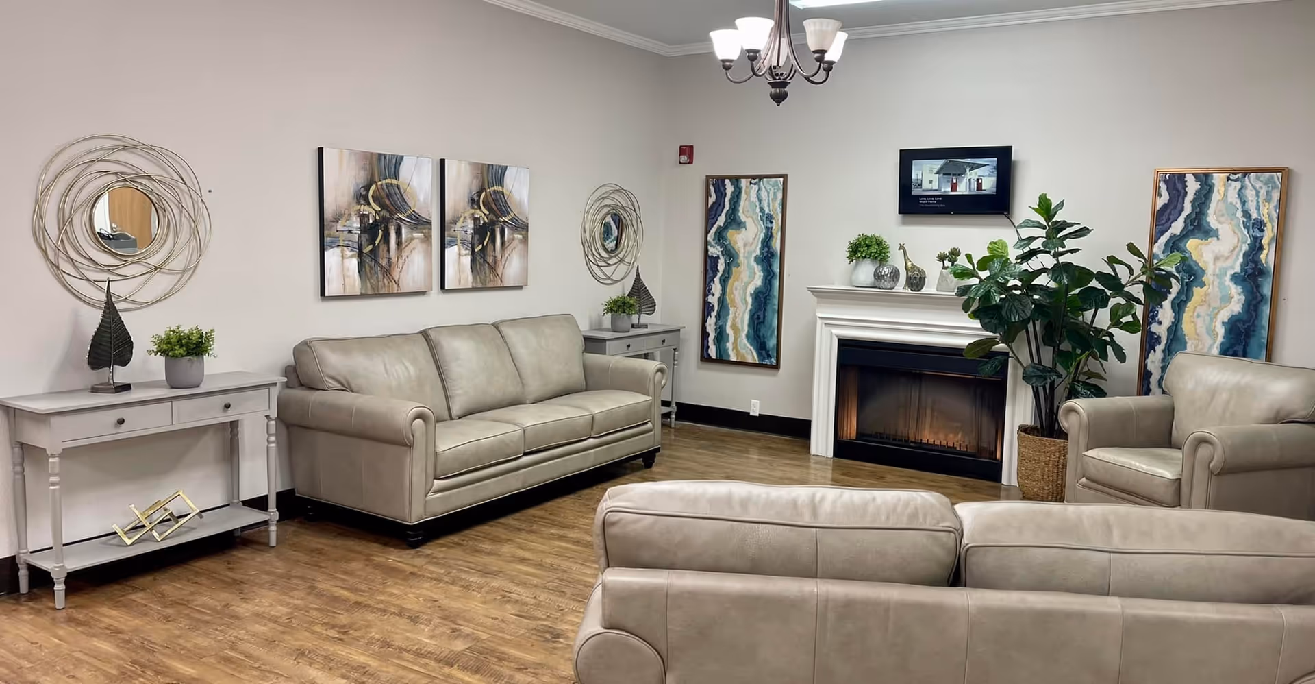 A spacious lounge area with beige leather seating around a fireplace, framed art, and potted plants in a senior living facility.