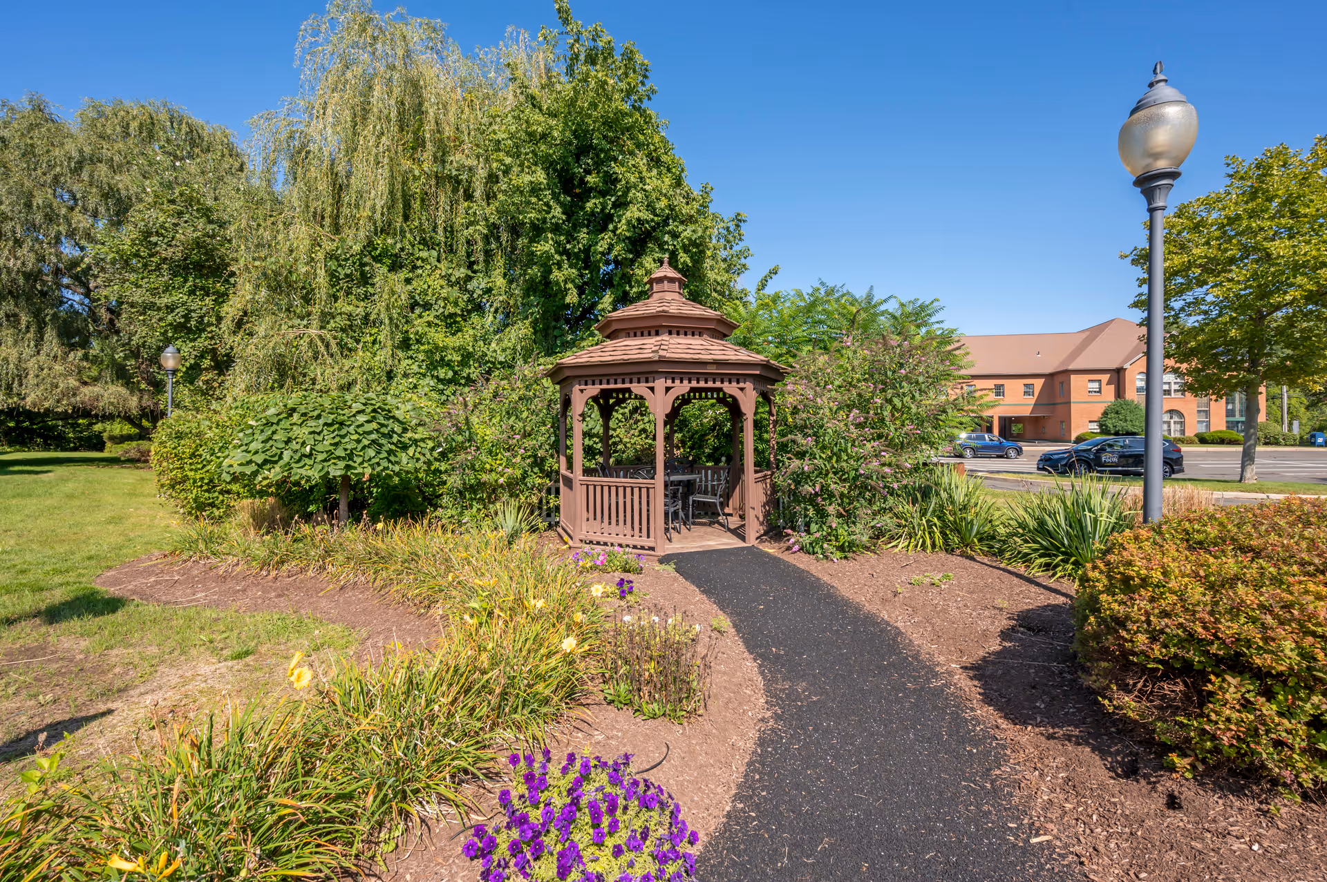 Wooden gazebo at the end of a paved path surrounded by landscaped garden beds, trees, and a building in the background.