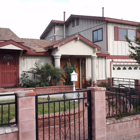 Front exterior view of a two-story residential building with a red door, a garage, and a small garden area enclosed by a brick and wrought iron fence.
