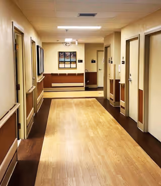 Well-lit interior hallway with wood flooring, handrails and multiple doors along the sides in a senior living facility.