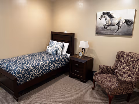 A small bedroom with a single bed, nightstand with a lamp and clock, a patterned armchair, and a horse painting on the wall.