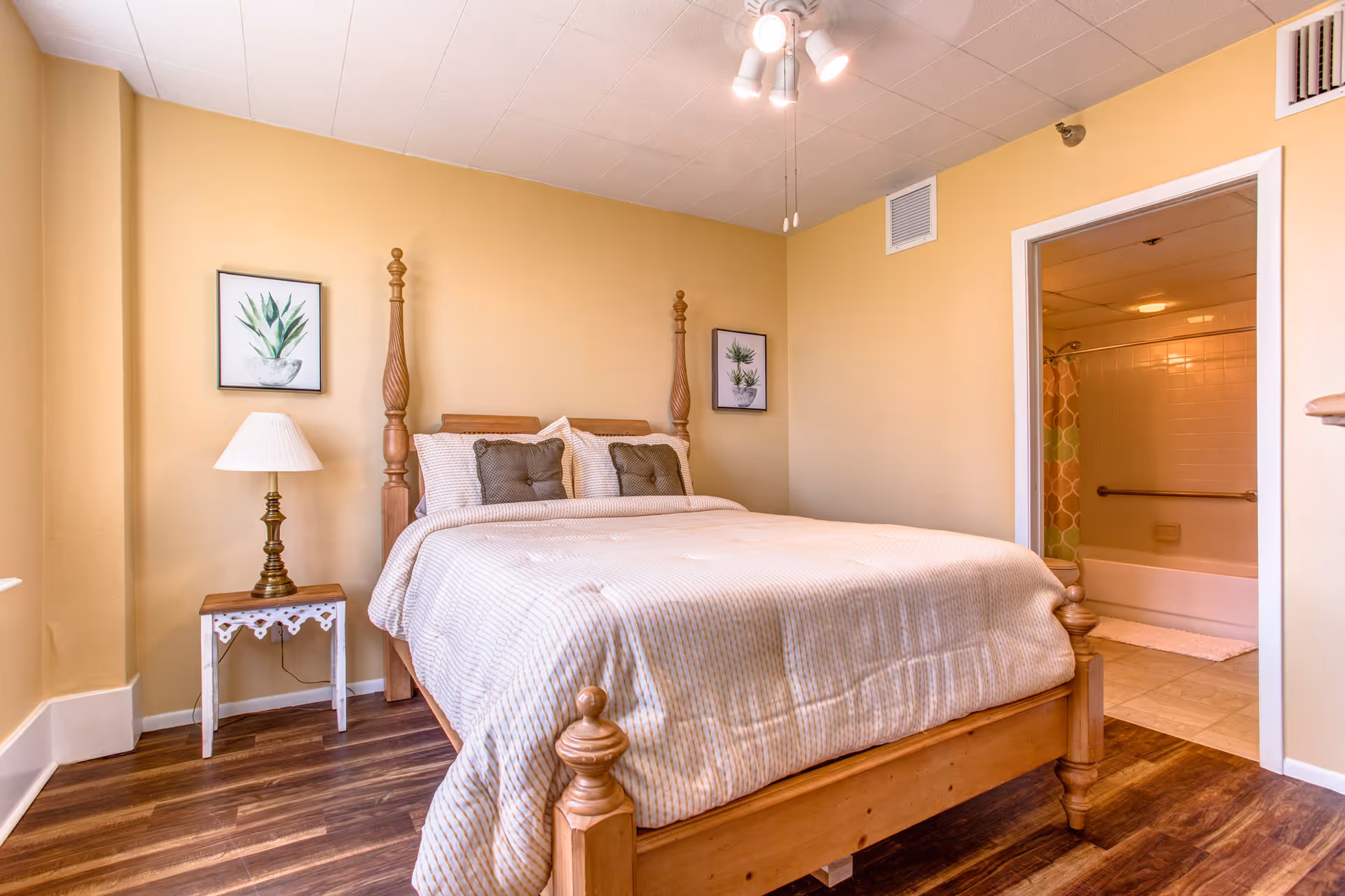 A cozy bedroom with a wooden four-poster bed made with beige striped bedding and two brown pillows. There is a small white side table with a brass lamp on the left side of the bed. Two framed botanical prints hang on the yellow walls above the bed. The room has wood flooring and a ceiling fan with lights. An open doorway on the right side reveals a bathroom with a bathtub and a colorful shower curtain.