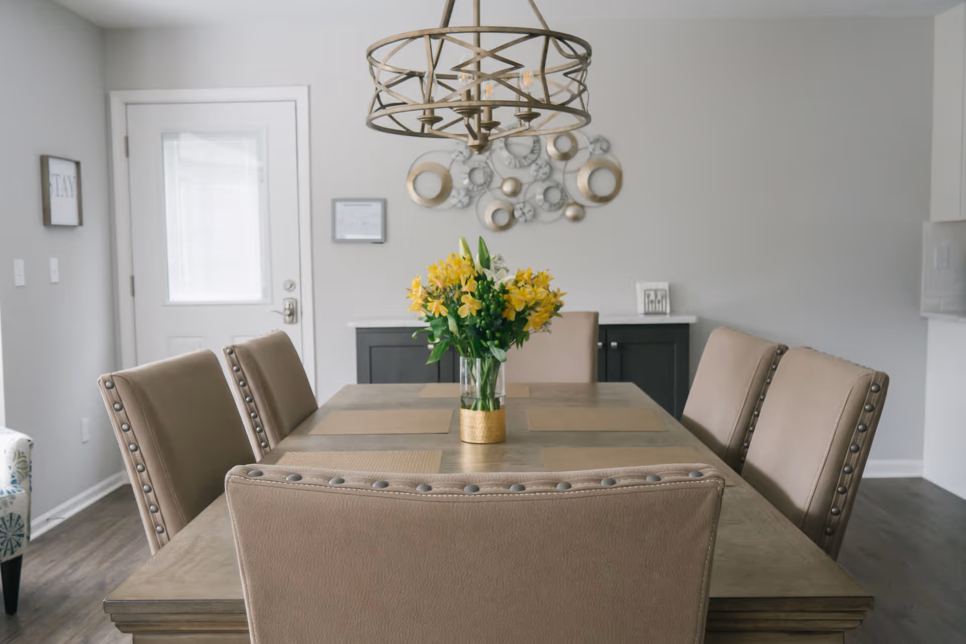 A modern dining room with a rectangular wooden table surrounded by six beige upholstered chairs. A vase with yellow flowers is centered on the table. A decorative metal wall art and a chandelier with a circular metal frame hang on the light gray wall. A white door with a window is visible in the background.