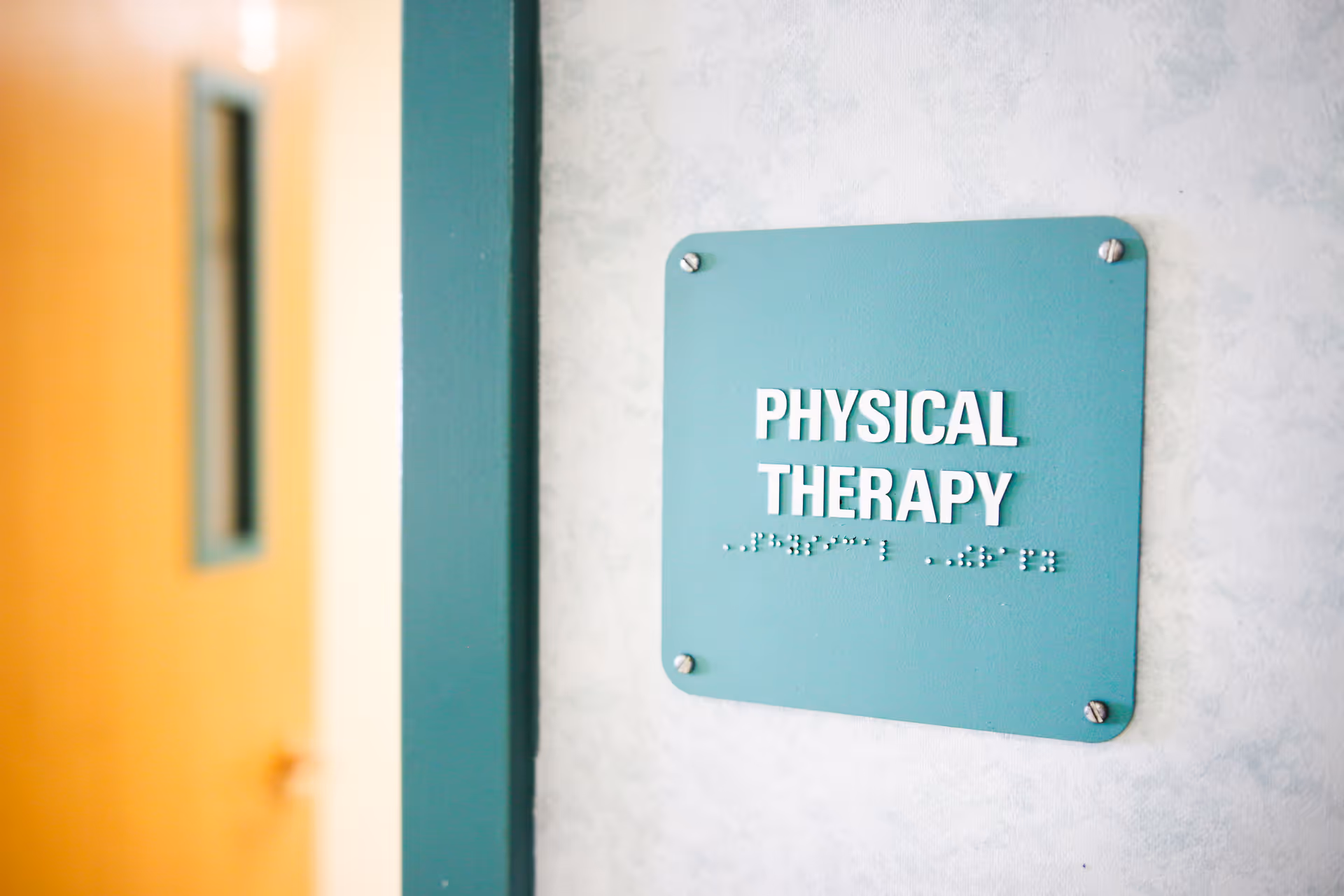 A teal sign mounted on a light-colored wall with the words 'PHYSICAL THERAPY' in white capital letters and Braille below. A wooden door with a window is partially visible to the left.