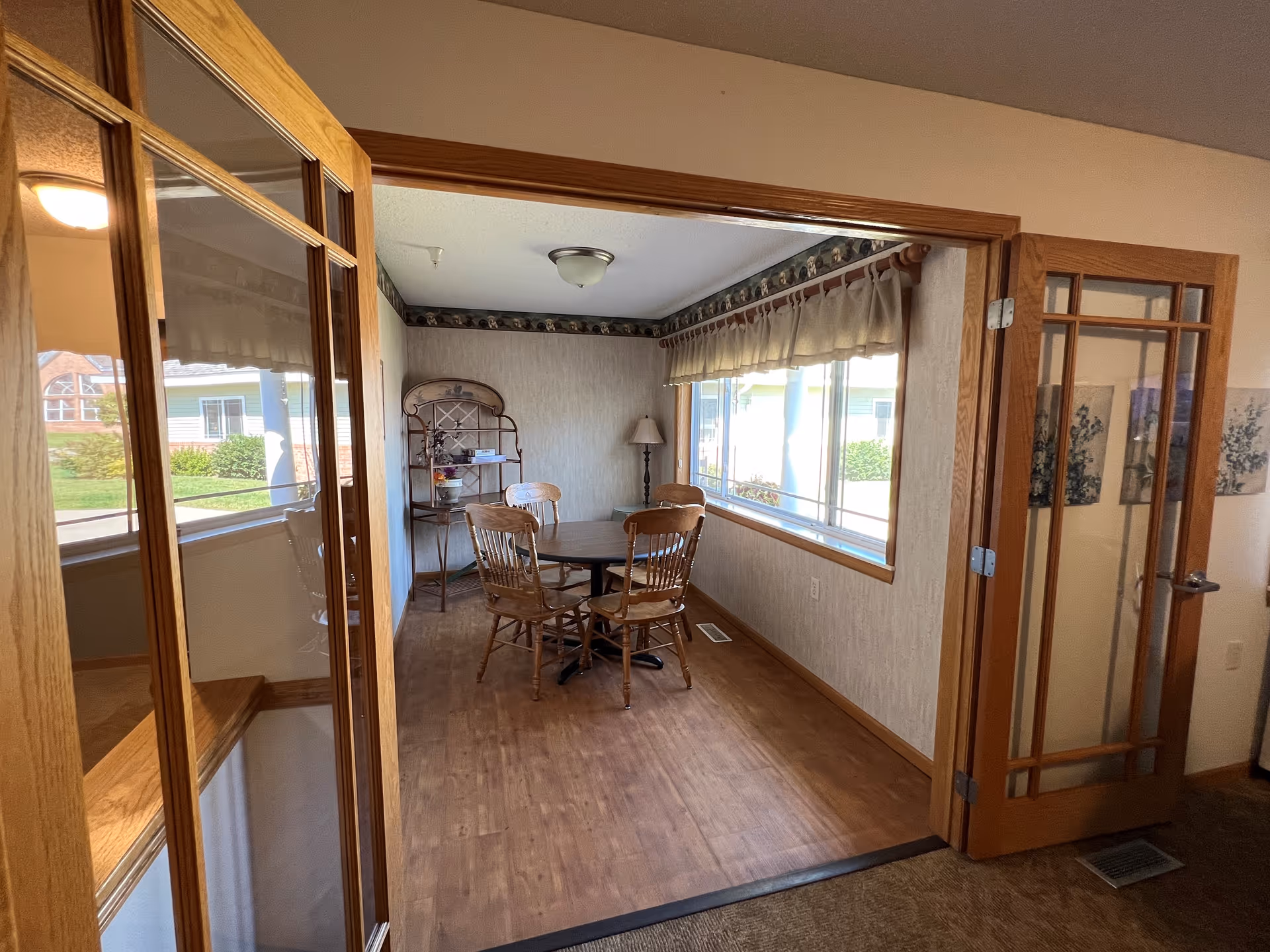 A small dining area with a round wooden table and four wooden chairs. The room has large windows with beige curtains allowing natural light to enter. There is a wooden shelving unit with decorative items and a floor lamp in the corner. The dining area is framed by wooden French doors with glass panels, and the floor is wood with a carpeted area outside the room.