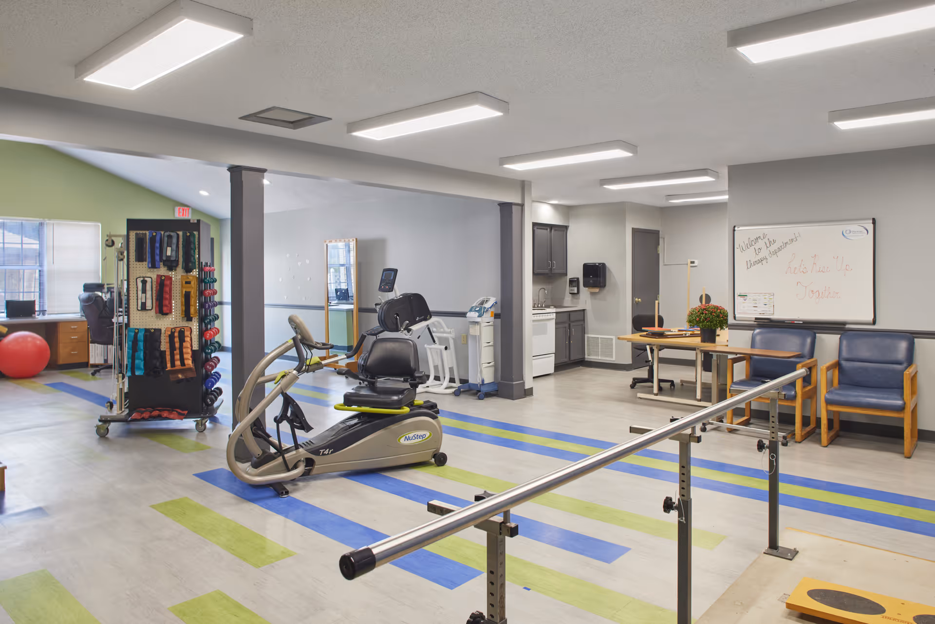A spacious physical therapy or rehabilitation room with exercise equipment including a NuStep recumbent cross trainer, parallel bars for walking support, a rack with various exercise bands and weights, and a small kitchenette area with cabinets and a sink. There are chairs and a table with a potted plant, and a whiteboard on the wall with a welcoming message.