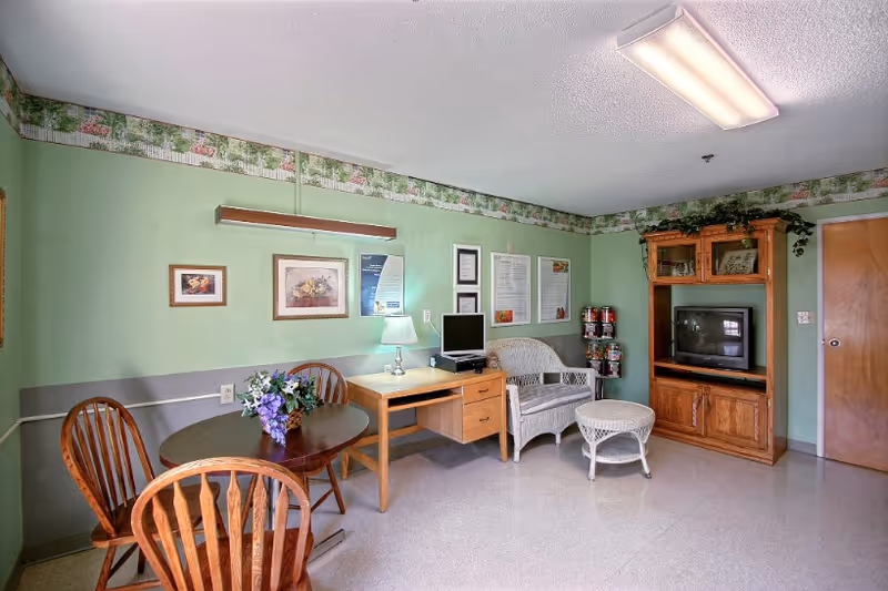 A cozy room with light green walls and floral border wallpaper. The room features a small round wooden table with three wooden chairs, a wooden desk with a lamp and a computer monitor, a white wicker loveseat and matching round wicker table, and a wooden entertainment center with a television. There are framed pictures and informational posters on the walls, and a closed wooden door on the right side.