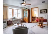 Bright living room in a retirement center with armchairs, a coffee table, a china cabinet and a small dining table visible through an open doorway.