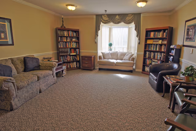 A cozy living room with a beige couch, two tall wooden bookshelves filled with books, a small wooden side table, a dark leather armchair, and a window with drapes letting in natural light.