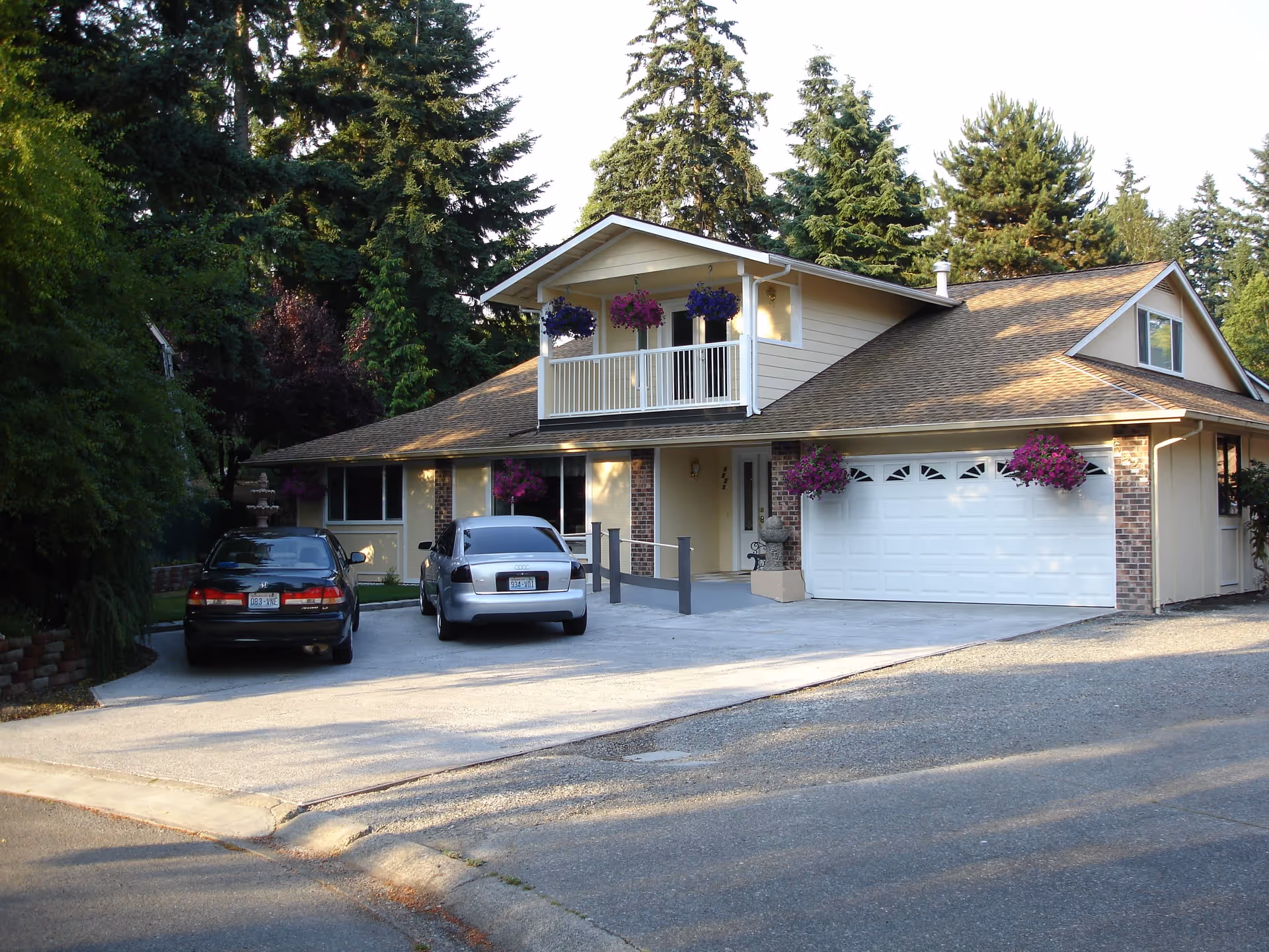 Exterior view of a two-story residential building with a sloped roof, a balcony with hanging flower pots, a garage door, and two cars parked in the driveway. The building is surrounded by tall trees and greenery.