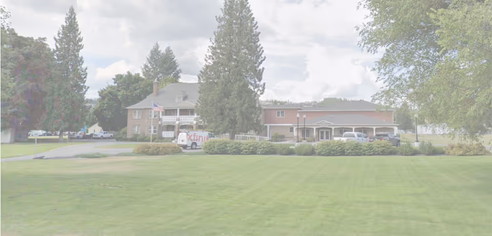 Wide view of Mallon Place facility showing a large green lawn in the foreground, with a two-story building and a single-story building behind it. Several tall trees and bushes surround the buildings, and a few vehicles are parked near the entrance. The sky is partly cloudy.
