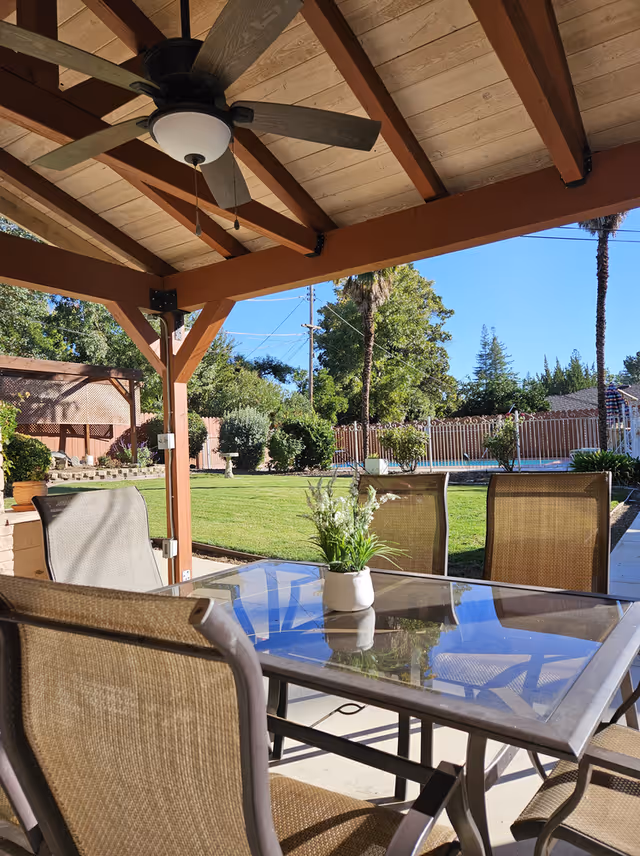 Outdoor patio area with a glass-top table and four mesh chairs under a wooden pergola with a ceiling fan. In the background, there is a well-maintained lawn, various trees, shrubs, and a fenced swimming pool under a clear blue sky.